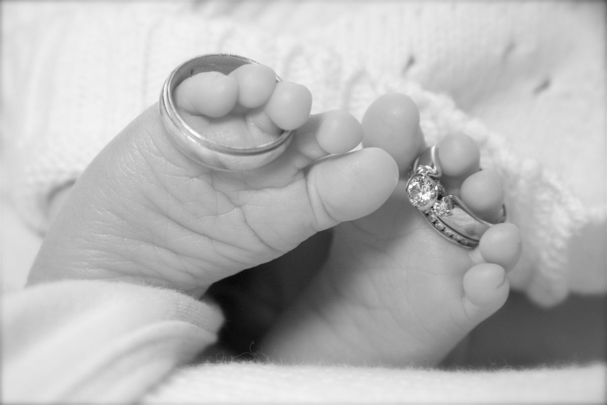 Intimate black and white photograph of newborn baby feet with parents' wedding rings resting on tiny toes, symbolizing generational connection and family blessing