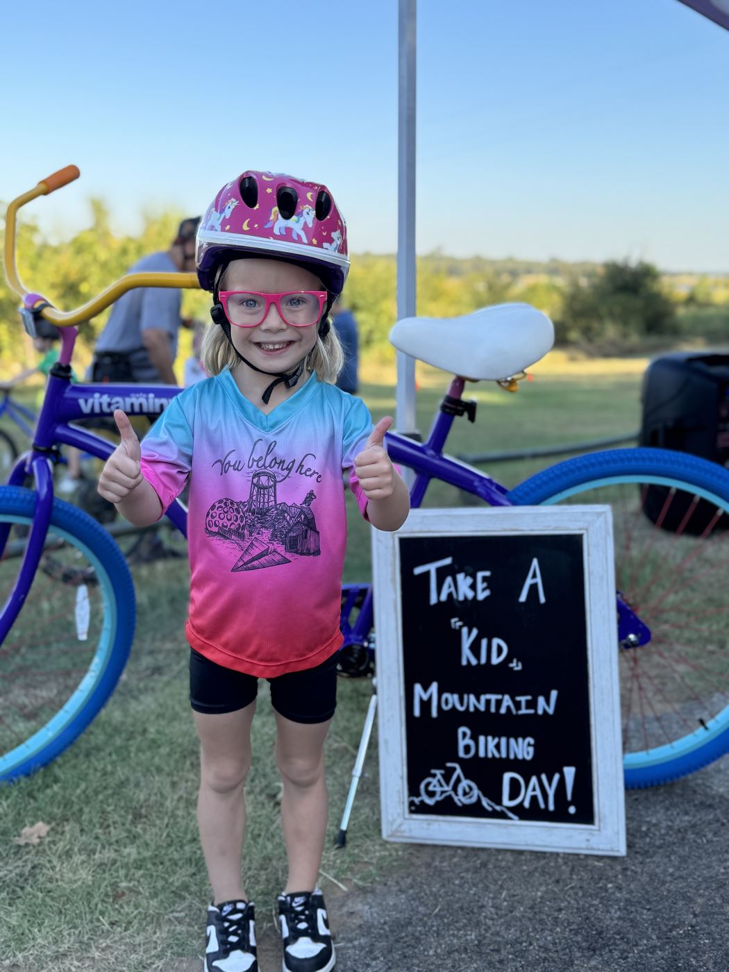 A young rider giving two thumbs up next to the Take a Kid Mountain Biking Day sign