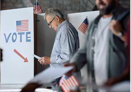 Man sits to review his ballot at a polling location before submitting his ballot. American flags and a Vote here sign with three American flags and man in the foreground walking to cast his ballot