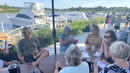 People socializing around a table next to a river with boats docked